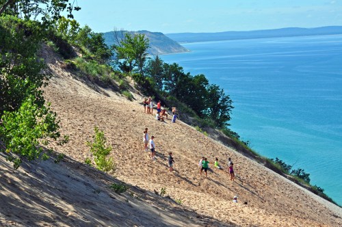 A sandy dune with people playing and exploring, overlooking a blue lake and lush green trees in the background.