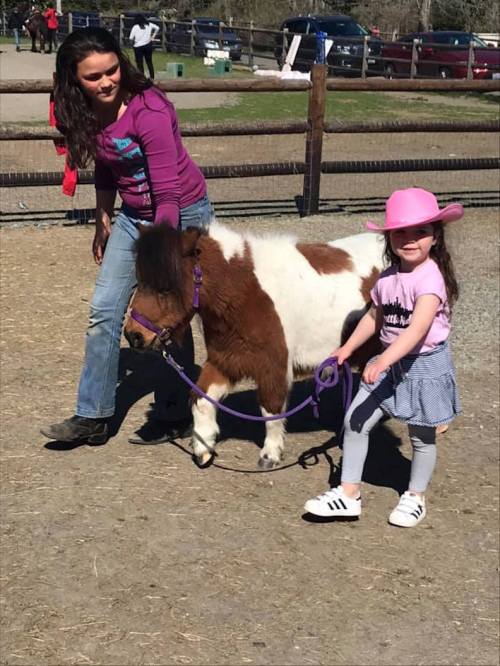 A girl in a pink hat walks a small pony with an adult nearby, both smiling in a sunny outdoor setting.