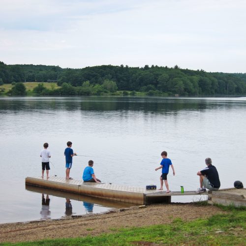 Five children on a dock by a calm lake, with trees in the background, engaged in fishing or playing.