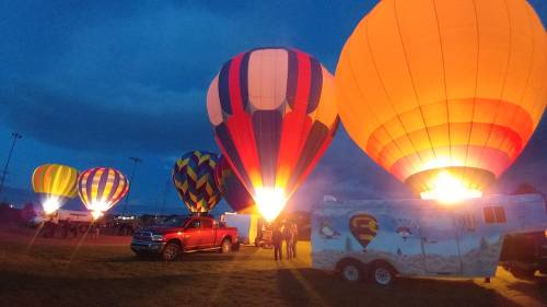 Colorful hot air balloons illuminated at night, with a truck and trailer nearby, set against a dark sky.