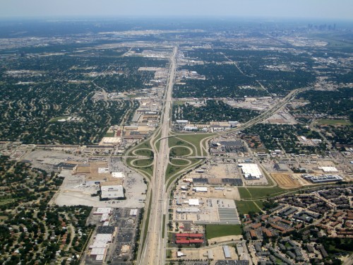 Aerial view of a highway interchange surrounded by urban development and green spaces. City skyline visible in the distance.
