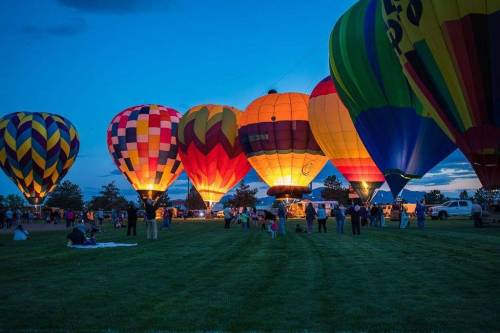 Colorful hot air balloons illuminated at dusk, with people gathered on a grassy field.
