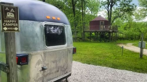 A vintage trailer parked near a treehouse in a green campsite, with a sign reading "Happy Camper Area."