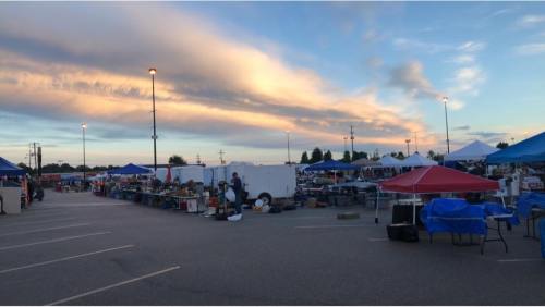 A bustling outdoor market at sunset, with colorful tents and vendors under a dramatic sky.