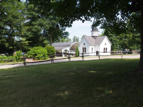 A small white church with a steeple surrounded by trees and a grassy area, featuring a wooden fence in the foreground.