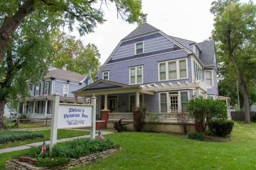 A large, historic purple inn with a sign, surrounded by greenery and American flags on a sunny day.