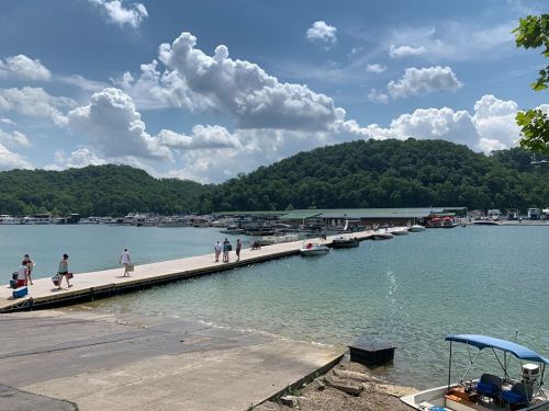 A scenic marina with boats docked, people walking on a pier, and lush green hills under a partly cloudy sky.