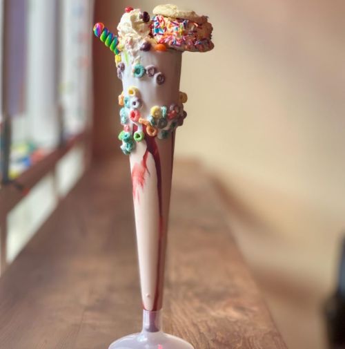 A tall milkshake topped with whipped cream, a donut, sprinkles, and colorful cereal, served in a fancy glass.