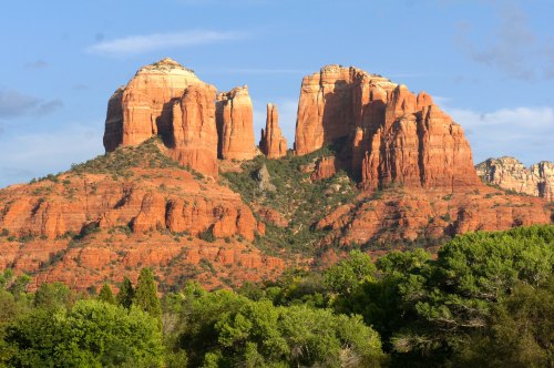 Red rock formations rise majestically against a blue sky, surrounded by lush green trees in a scenic landscape.