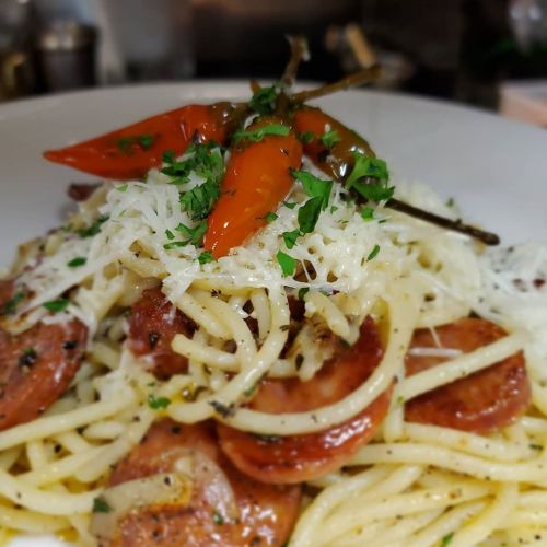 A plate of spaghetti topped with sliced sausage, grated cheese, and garnished with peppers and herbs.