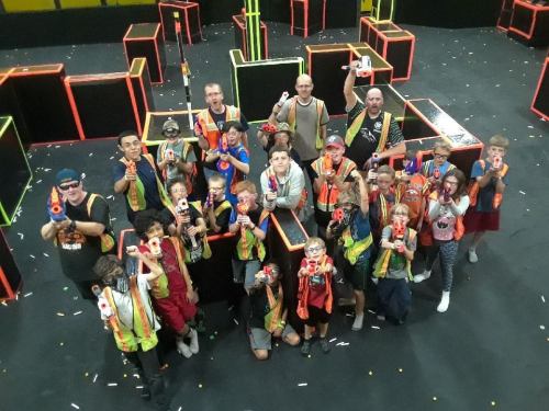 A group of children and adults in colorful vests, holding toy blasters, posing in a vibrant indoor laser tag arena.