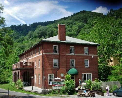 A brick building with a green awning surrounded by trees and mountains, featuring a welcoming sign and outdoor seating.