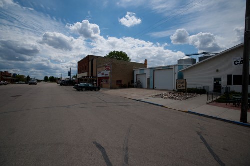 A quiet street scene featuring a cafe, shops, and parked cars under a partly cloudy sky.