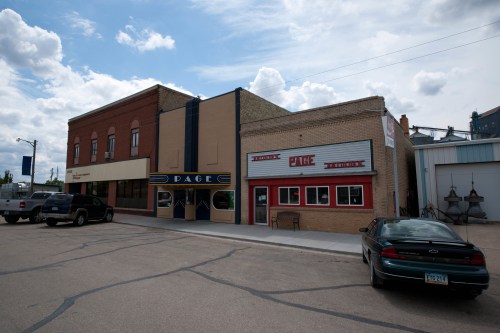 A street view of a small town featuring brick buildings, a theater, and a hardware store under a cloudy sky.