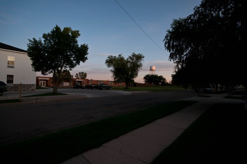 A quiet street at dusk, featuring trees, a water tower in the distance, and a few parked cars.