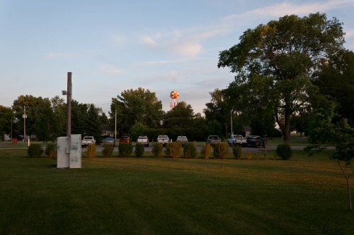A grassy area with trees and parked cars, featuring a distant tower illuminated by the setting sun.
