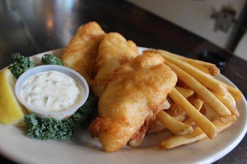A plate of golden fried fish fillets with French fries, tartar sauce, a lemon wedge, and a garnish of kale.
