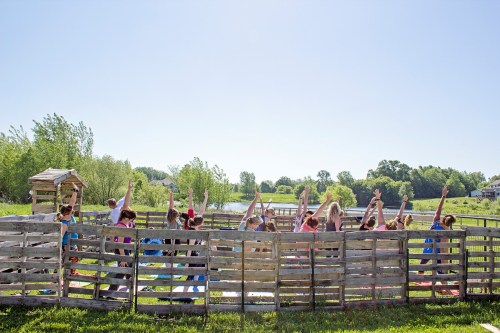A group of children outdoors, raising their hands in a fenced area with trees and a pond in the background.