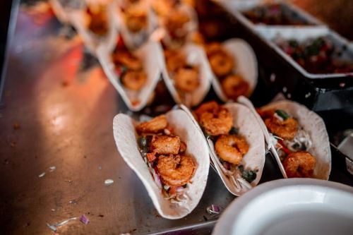 A close-up of shrimp tacos on a metal tray, garnished with vegetables and served in soft tortillas.