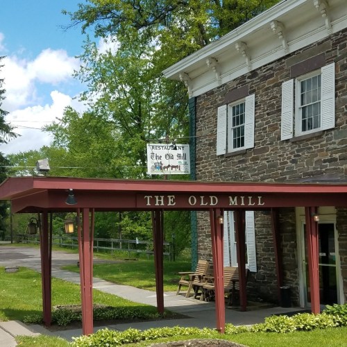A stone building with a red awning displaying "The Old Mill" sign, surrounded by greenery and blue skies.