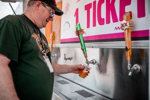 A man in a green shirt pours a drink from a colorful tap at a festival booth.