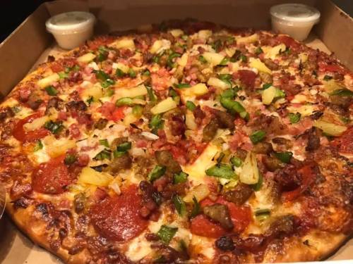 A pizza topped with pepperoni, green peppers, pineapple, bacon, and sausage, served in a pizza box with two dipping sauces.