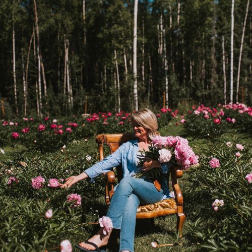 A woman sits in an ornate chair among blooming peonies, holding a bouquet and enjoying the vibrant garden.