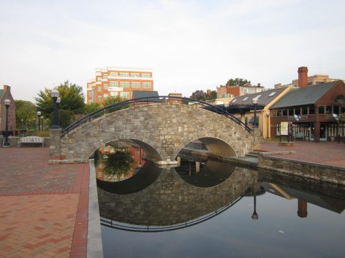 A stone bridge arches over a calm canal, reflecting nearby buildings and trees in the water.