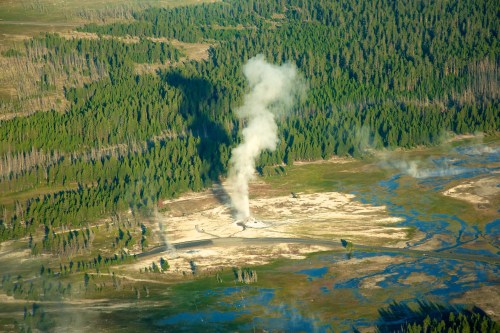 Aerial view of a geothermal area with steam rising from the ground, surrounded by green forests and water features.