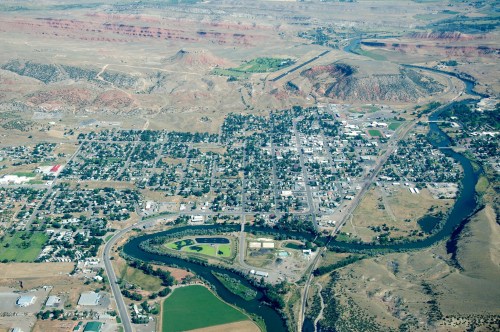 Aerial view of a small town surrounded by hills and a river, with green fields and roads visible.