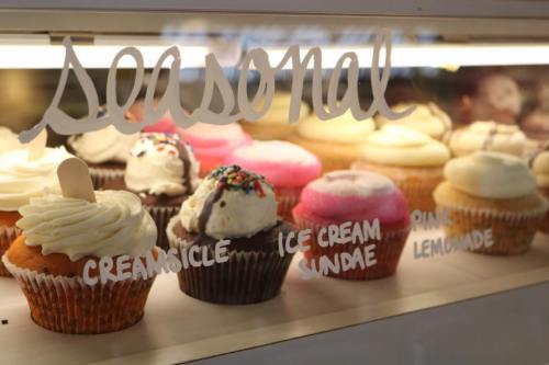 A display of colorful cupcakes with various toppings, labeled "seasonal" in a bakery setting.