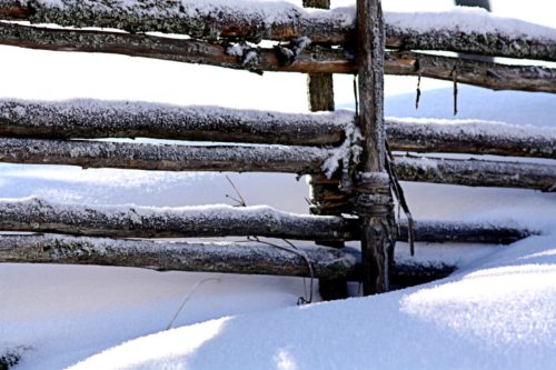 A rustic wooden fence covered in snow, with a soft white blanket of snow on the ground.