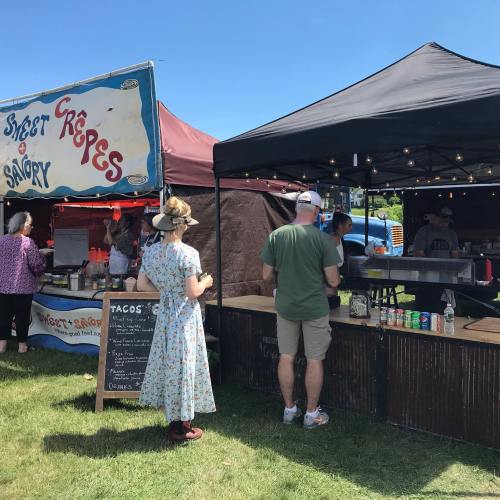 A food market scene with a woman in a floral dress and two people ordering at taco and crepe stalls under a blue sky.