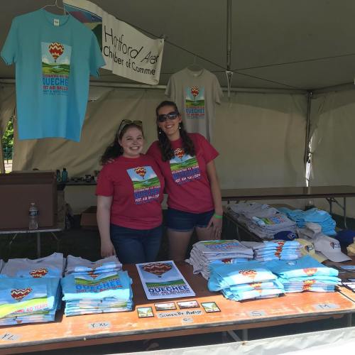 Two women in colorful t-shirts stand behind a table filled with various t-shirts and promotional materials at a fair booth.
