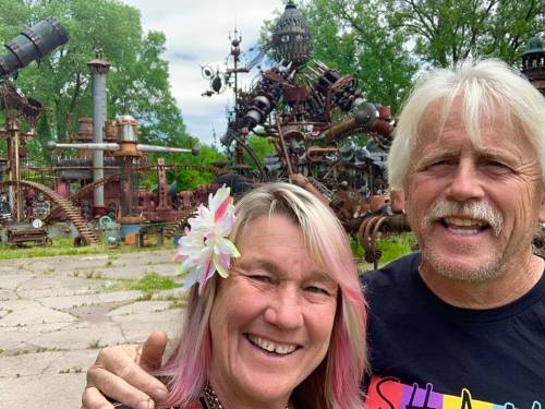 A smiling couple poses for a selfie in front of a whimsical, steampunk-inspired sculpture garden.