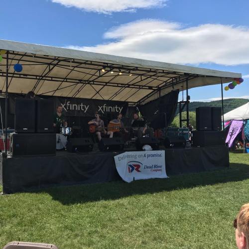 A band performs on stage at an outdoor event, with colorful decorations and a clear blue sky in the background.