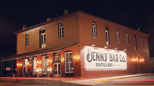A brick distillery building at night, featuring a large sign that reads "Jenny Bar Co. Distillery." Warm lights illuminate the facade.