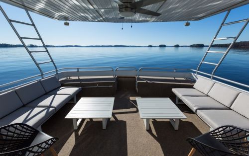 A spacious boat deck with gray seating and white tables, overlooking a calm lake and clear blue sky.