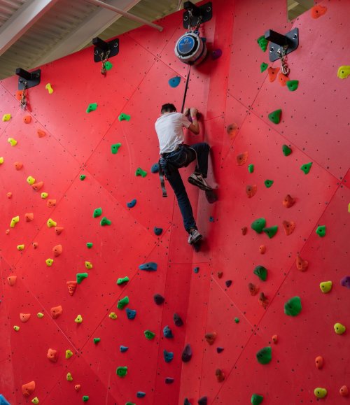 A child climbs a colorful indoor rock wall, reaching for holds on a bright red surface.