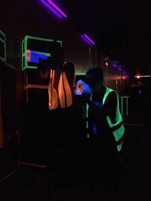 Two people in neon vests aim laser tag guns in a dark, glowing arena with colorful lights.