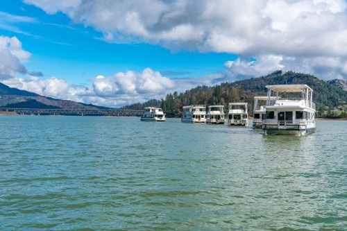 A serene lake scene with houseboats anchored, surrounded by green hills and a blue sky with fluffy clouds.