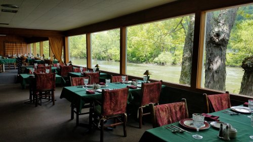 A cozy restaurant with green tables and chairs, overlooking a serene river and lush greenery through large windows.