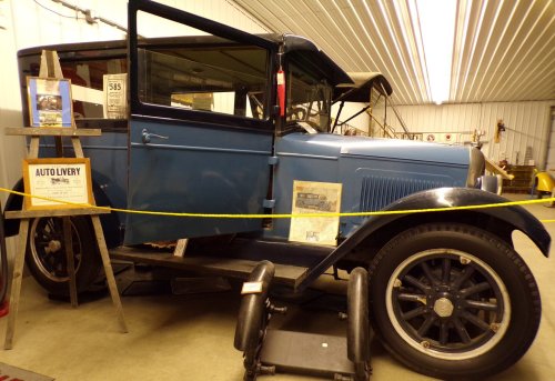 A vintage blue car displayed in a museum, with informational signs and a yellow rope barrier.