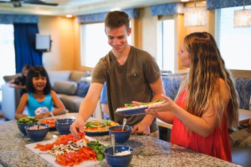 A young man and woman serve food at a kitchen counter, while a girl smiles in the background. Colorful dishes are displayed.