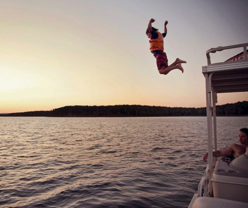 A child in a life jacket jumps off a boat into a lake at sunset, while another person relaxes on the boat.
