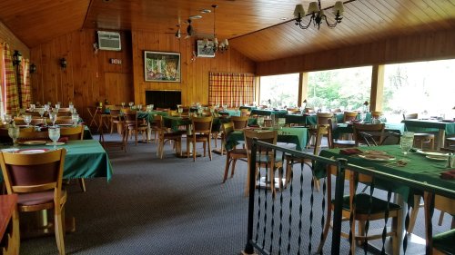 A spacious dining room with wooden walls, green tablecloths, and neatly set tables, surrounded by large windows.