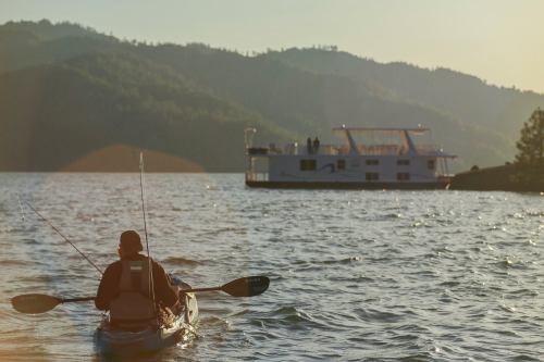 A person in a kayak fishing near a houseboat on a calm lake, surrounded by mountains at sunset.