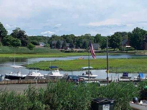 A peaceful waterfront scene with boats docked, an American flag, and lush greenery under a partly cloudy sky.