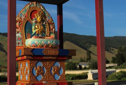 A colorful statue of a deity stands on a decorated pedestal, surrounded by mountains and a clear blue sky.
