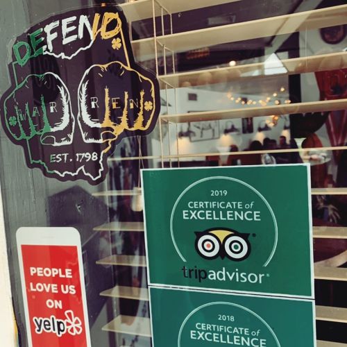 A window display featuring a "Defend Warren" sticker and TripAdvisor certificates of excellence from 2018 and 2019.